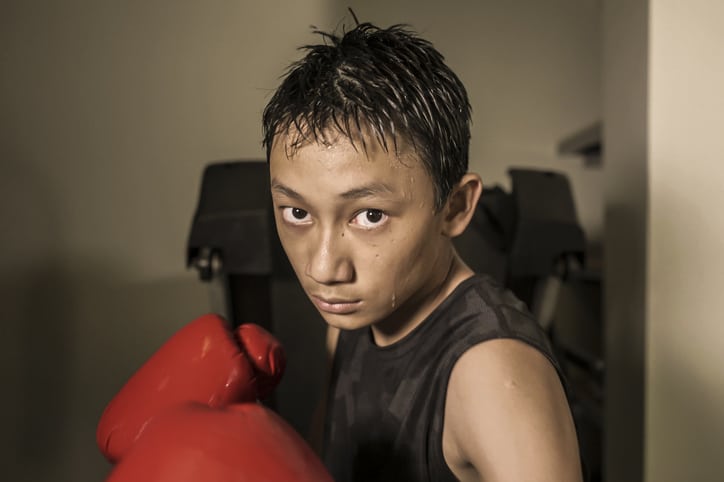 tough and cool young boy punching on heavy bag . 13 or 14 years old Asian teenager training Thai boxing workout looking defiant as a badass fighter practicing sport at fitness club