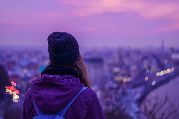 Hipster girl traveler with backpack looking at winter evening cityscape, purple violet sky and blurred city lights