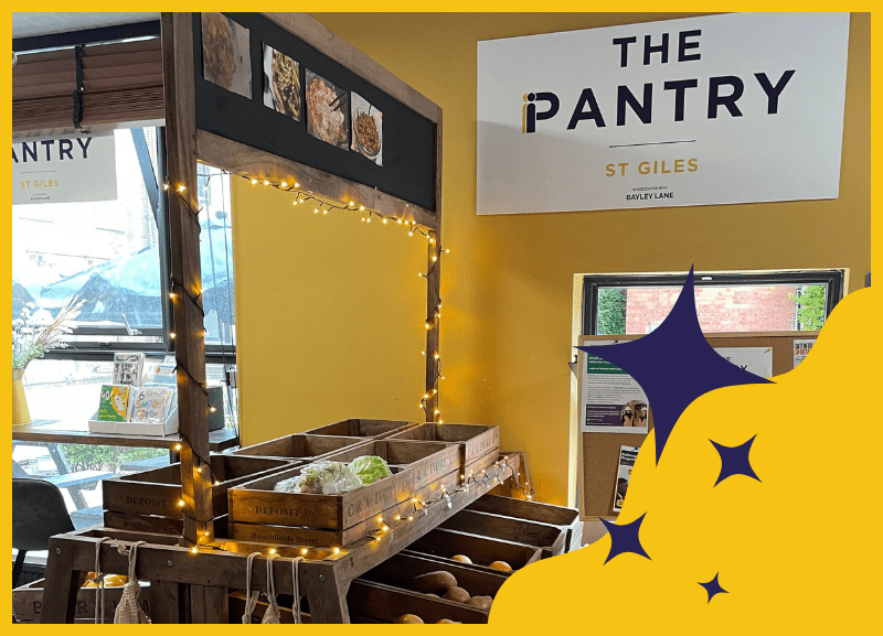 Photograph of The Pantry decorated for Christmas with fresh produce and the St Giles Trust Pantry logo in the top right corner. Bottom right corner has a graphic of blue stars over a wavey yellow shape.
