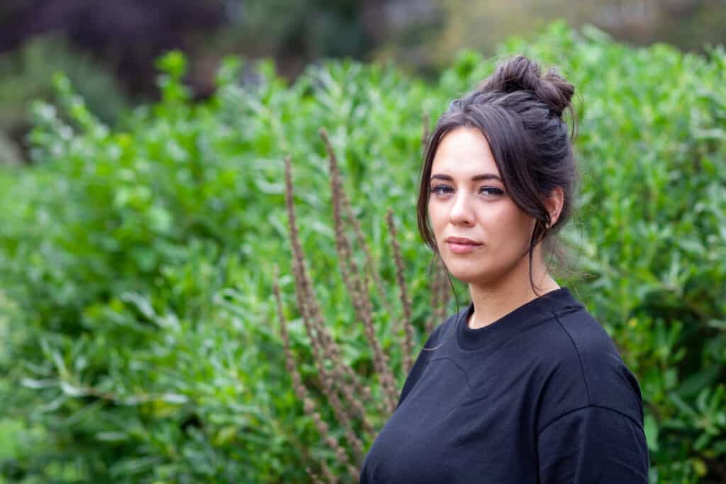 Photograph of Caitlin looking at the camera. She is stood in front of a hedge wearing a black T-Shirt.