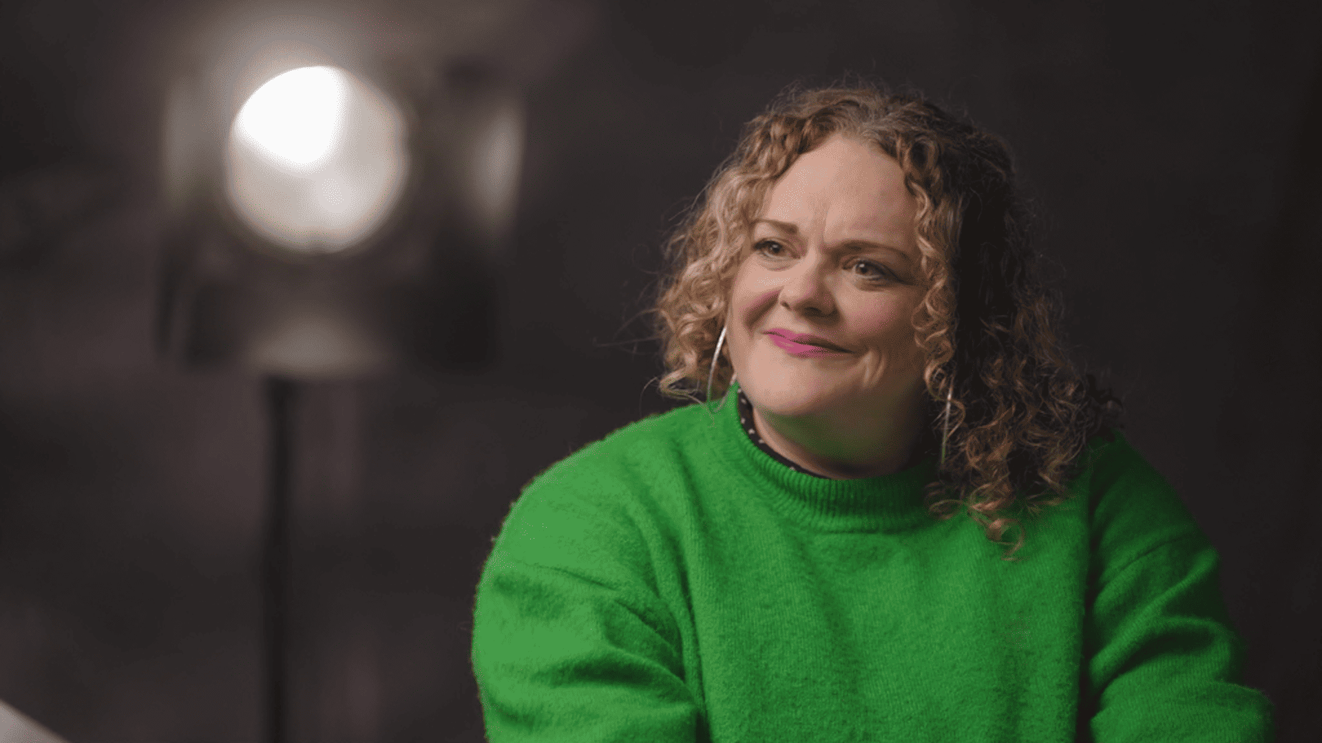 Photograph of woman wearing a green jumper and smiling with a large lamp behind her.