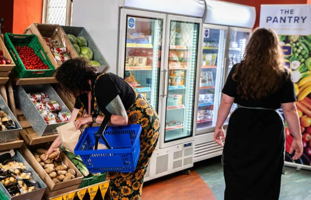 Photograph inside ST Giles Pantry Norwich. Woman in dungarees picking fresh produce from shelves next to fridges full of high quality produce.