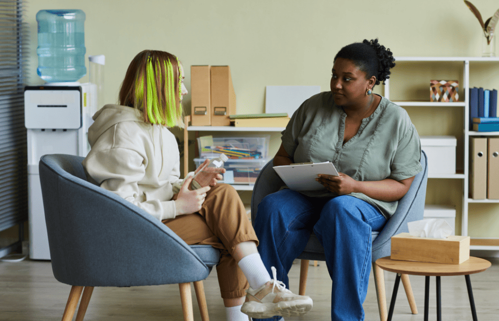 A young black female listens to a white teenage girl with coloured hair. They are sitting in armchairs in a office.