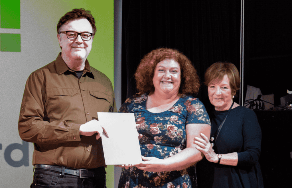 Photograph of Carly and Adam from St Giles East Of England Pantry Team smiling and being awarded a Certificate on stage with Delia Smith.