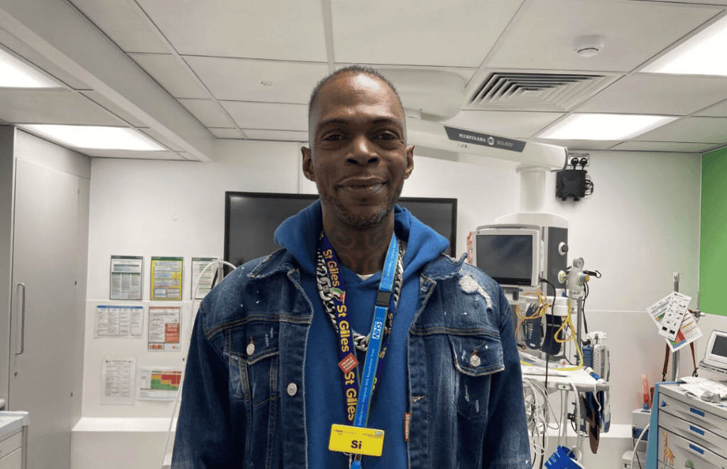Photograph of Si Philbert stood in a hospital room with machinery behind him. He is smiling and wearing a St Giles lanyard.