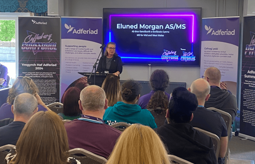 WOman stood at front of conference room, presenting to full room. Screen behind her says "Eluned Morgan - AS/MS for Mid and West Wales."