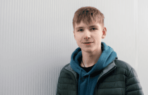 Young boy looking in to camera wearing a padded coat, stood in front of a corrugated mental sign.