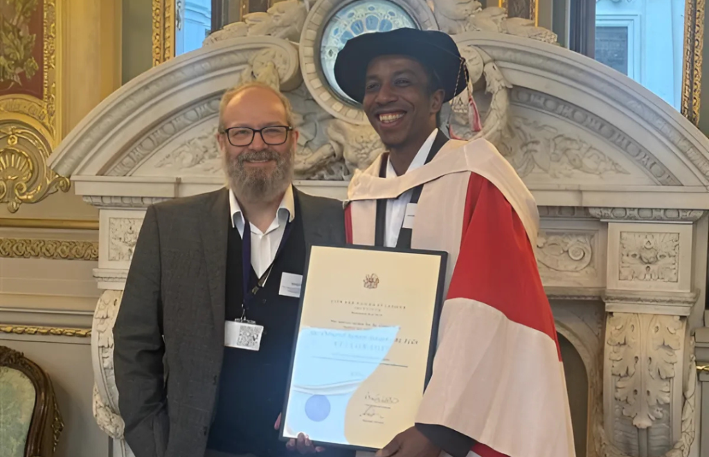 Photograph of Junior Smart holding a framed certificate while wearing formal and ornate robes and a hat. Junior is stood next to Andy Cross, Executive Director of Services at St Giles Trust.