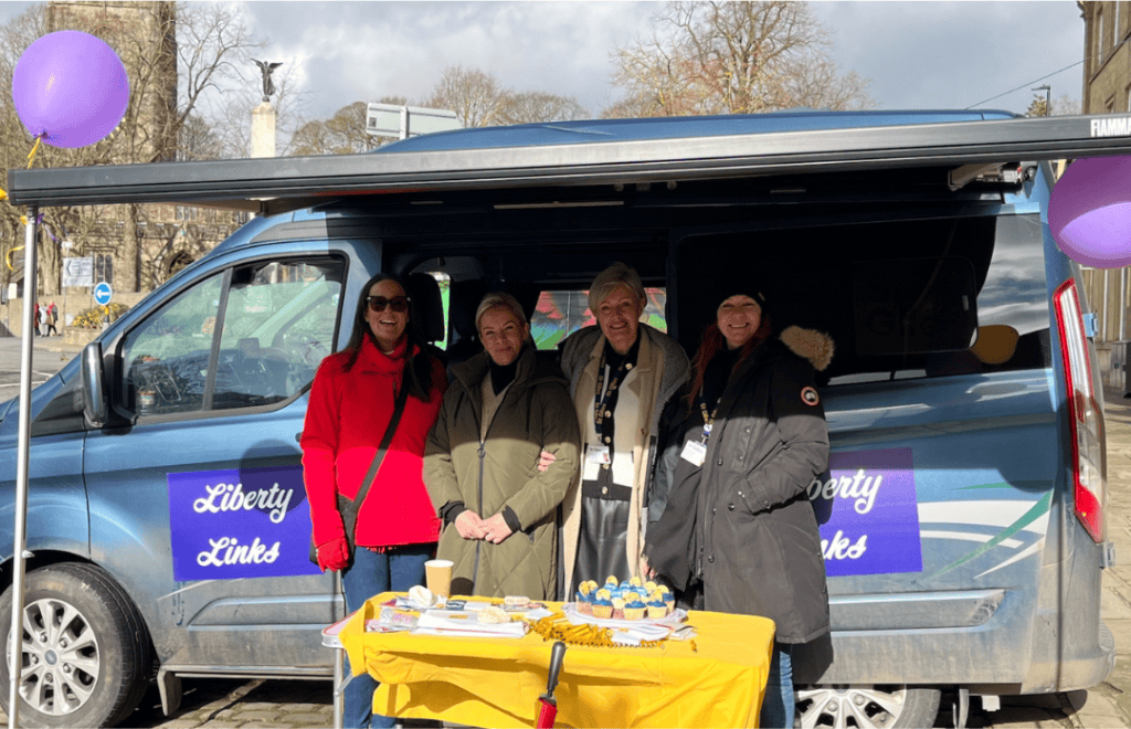 4 women stood behind a table with a yellow table cloth on it. They are infront of a van with dark blue branding 'Liberty Links' on the doors and side.