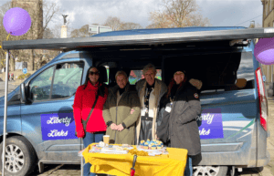4 women stood behind a table with a yellow table cloth on it. They are infront of a van with dark blue branding 'Liberty Links' on the doors and side.