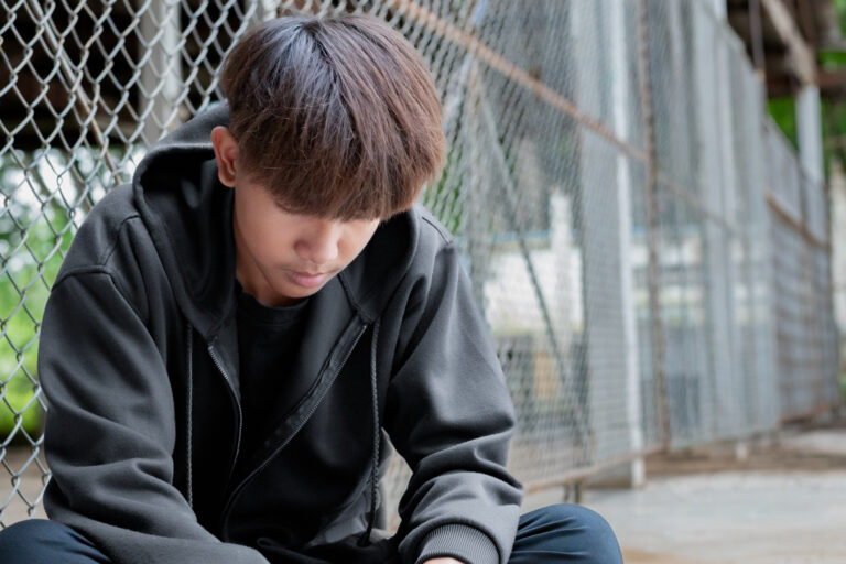 Asian preteen boy in a black hoodie sits with his knees pressed against a metal fence panel in a juvenile detention facility, awaiting further release, freedom and detention of people concept.