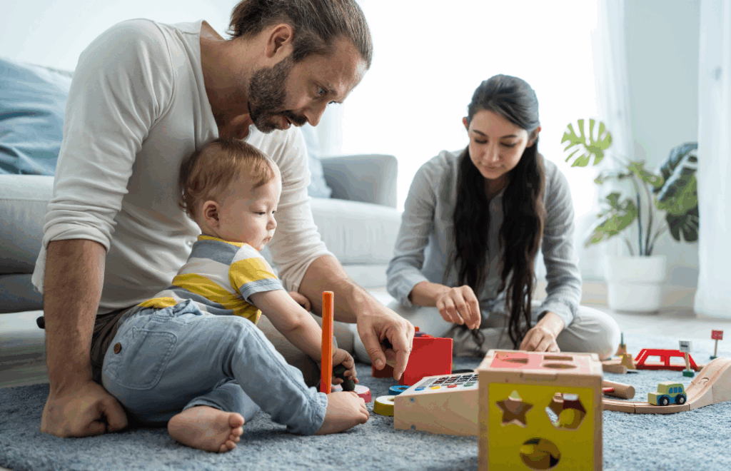 New parents sat with the baby on the carpet in a bright room, playing with toys.