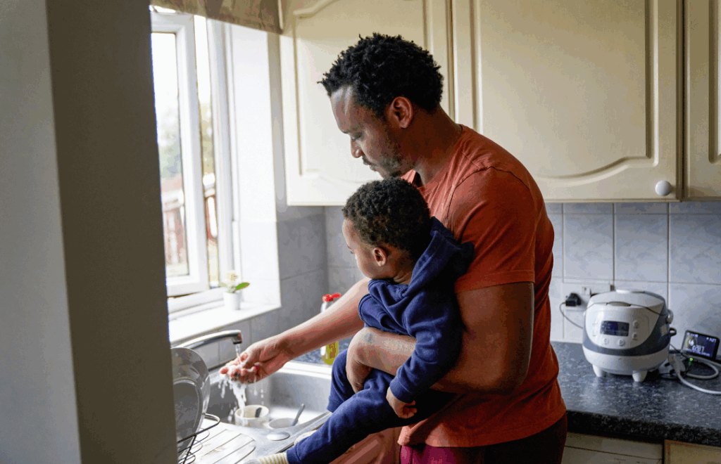 Dad holding a baby stood in front of a sink, running the tap and washing his hands.