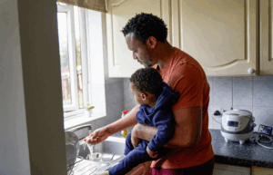 Dad holding a baby stood in front of a sink, running the tap and washing his hands.