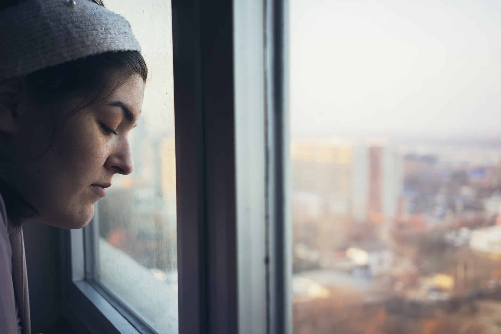 Half profile of a young girl looking at the city through a room window.