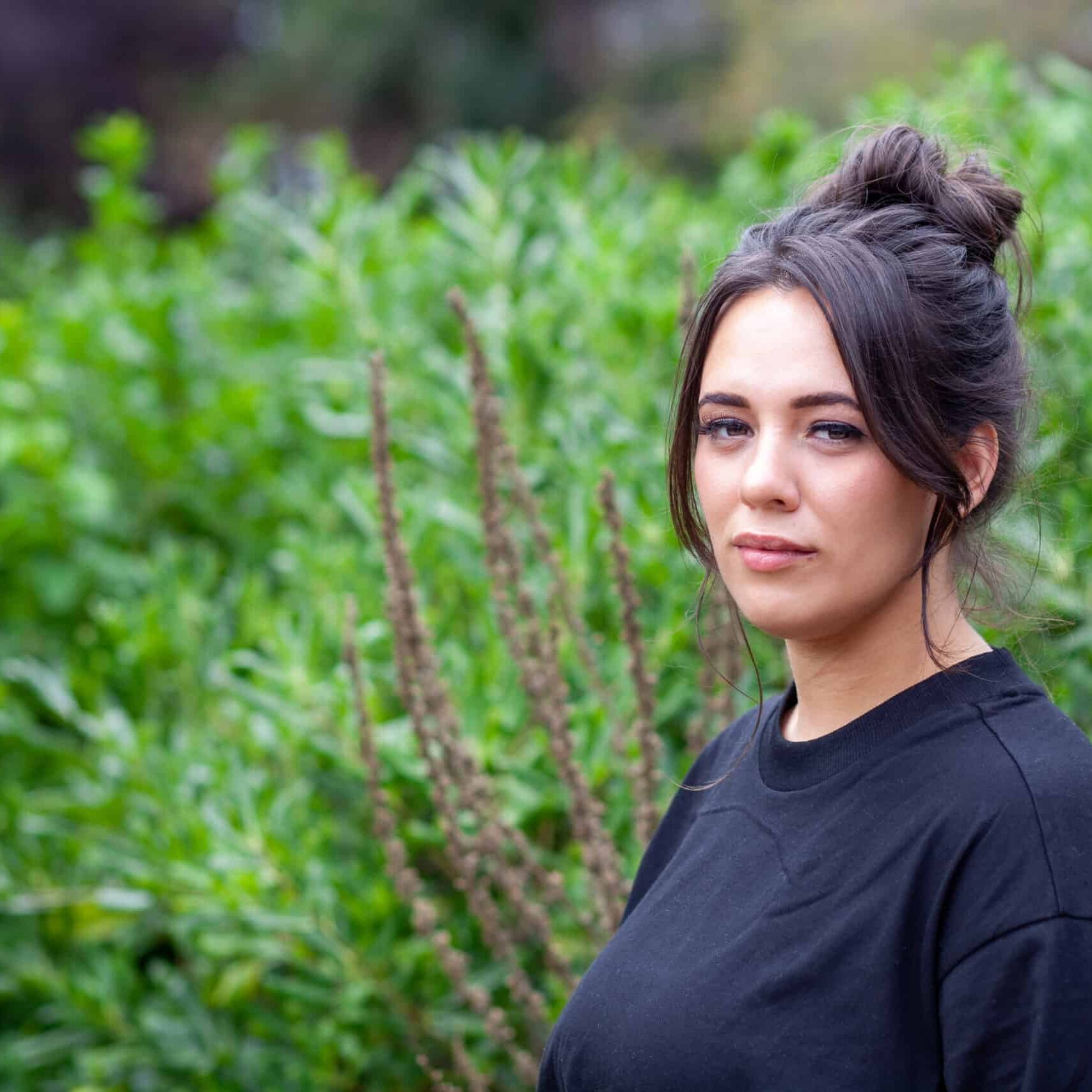 Photograph of Caitlin looking at the camera. She is stood in front of a hedge wearing a black T-Shirt.