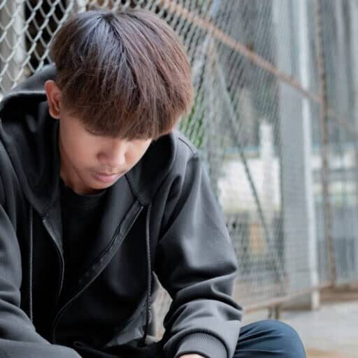 Asian preteen boy in a black hoodie sits with his knees pressed against a metal fence panel in a juvenile detention facility, awaiting further release, freedom and detention of people concept.