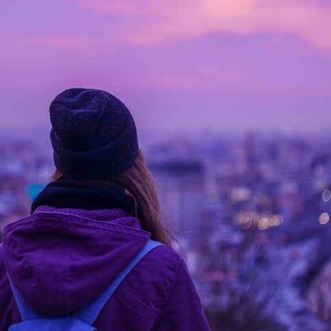 Hipster girl traveler with backpack looking at winter evening cityscape, purple violet sky and blurred city lights